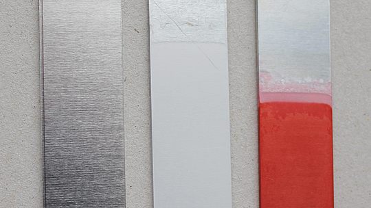 3 auf unterschiedliche Art behandelte Metallstreifen, links silber-metallisch glänzend, Mitte weißlich-matt, rechts an der Oberseite metallisch, in den unteren 2/3 rot