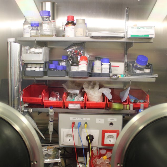 Sicht auf das innere einer Glovebox (Handschuhkasten), an der Rückwand Kisten und Gläser mit Versuchsmaterialien
